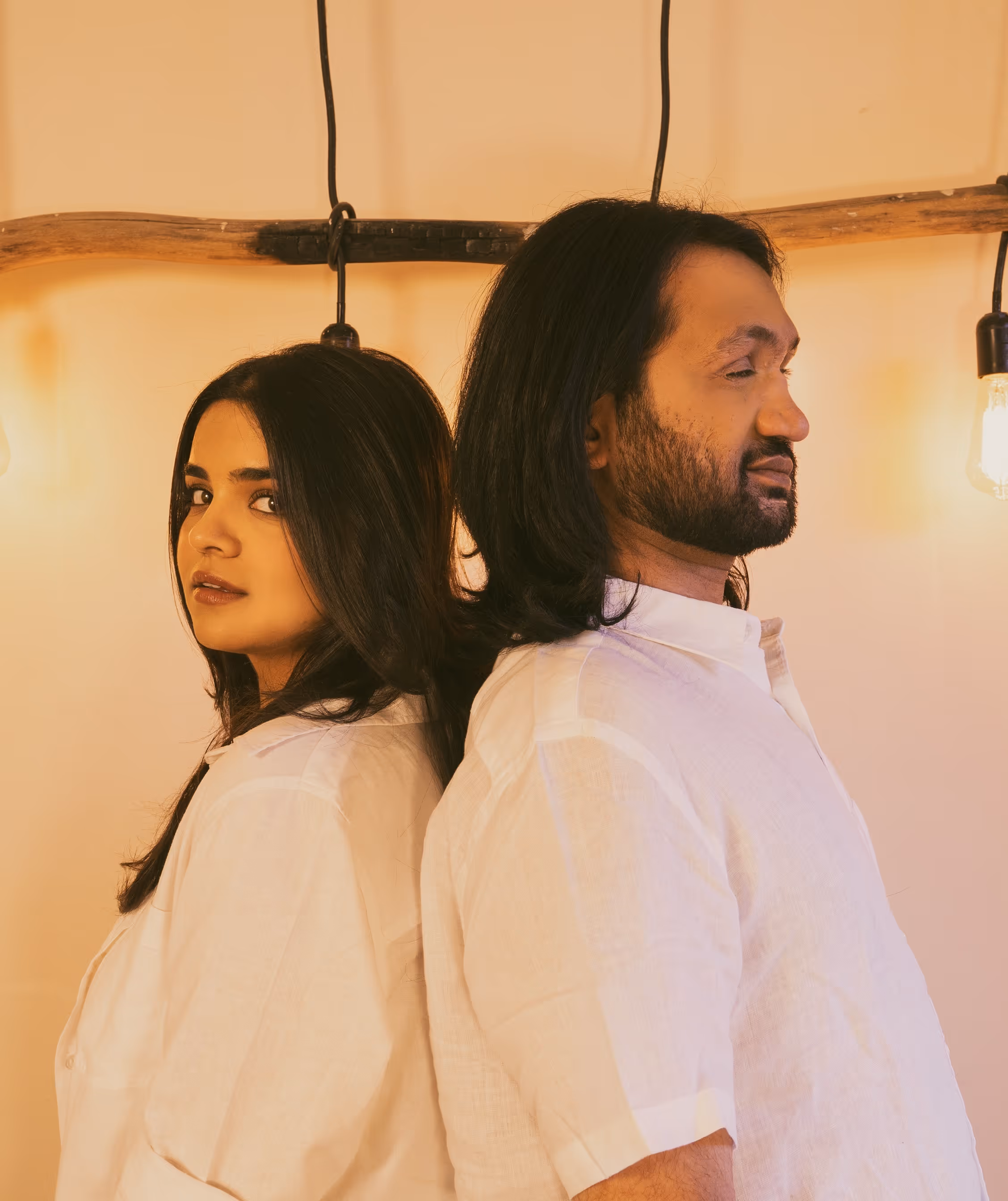 Eshaan Sood and Priyaana stand back-to-back under warm lighting, both dressed in coordinated white outfits. They face opposite directions with composed expressions. Vintage-style hanging bulbs and a raw wooden beam provide a minimalist, aesthetic backdrop.