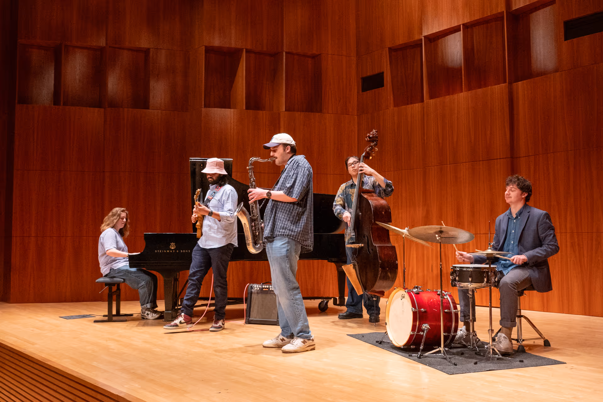 Eshaan Sood and the Sonic Alchemists perform with relaxed joy on a wooden stage. He stands near a grand piano and an upright bass, while a saxophonist and drummer complete the ensemble. Everyone appears to be enjoying the groove, with visible smiles and interaction